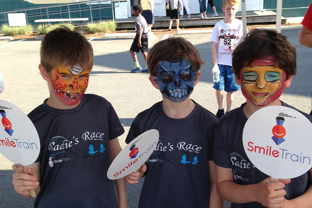 Three boys with facepaint smiling and holding Smile Train signs at Sadie's Race