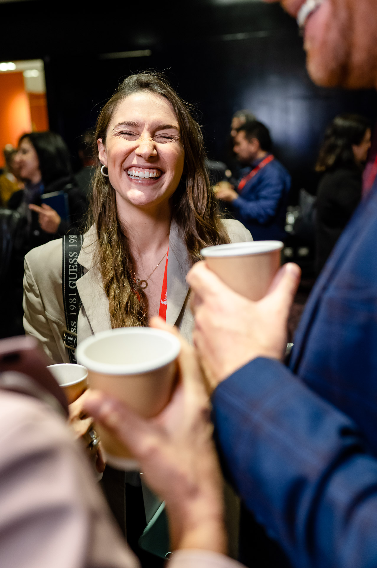 Camile Richards in a crowded room smiling. Two people in the foreground are holding coffee cups and out of focus