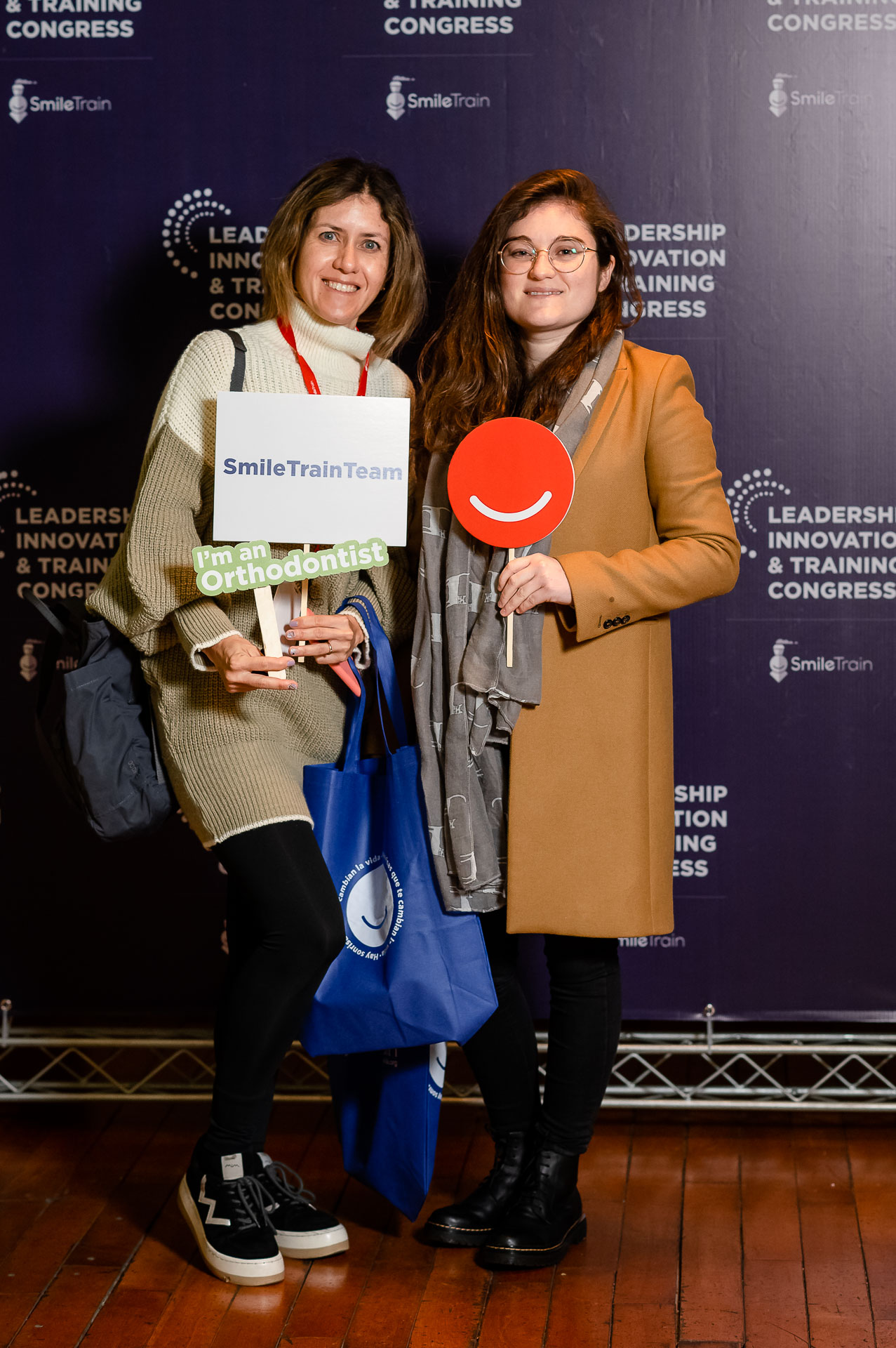 Francisca Salazar and Scarlette Norambuena posing with photobooth props. On the left, a sign reading ‘Smile Train Team’ and ‘I’m an Orthodontist.’ On the right, the orange smile logo