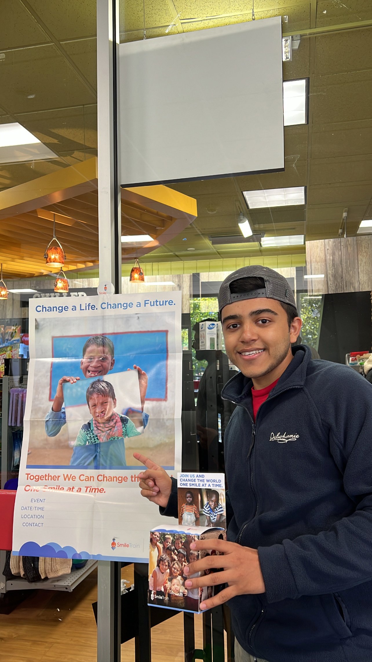 A member of Project Dental All holding a Smile Train money box and pointing to a Smile Train event poster