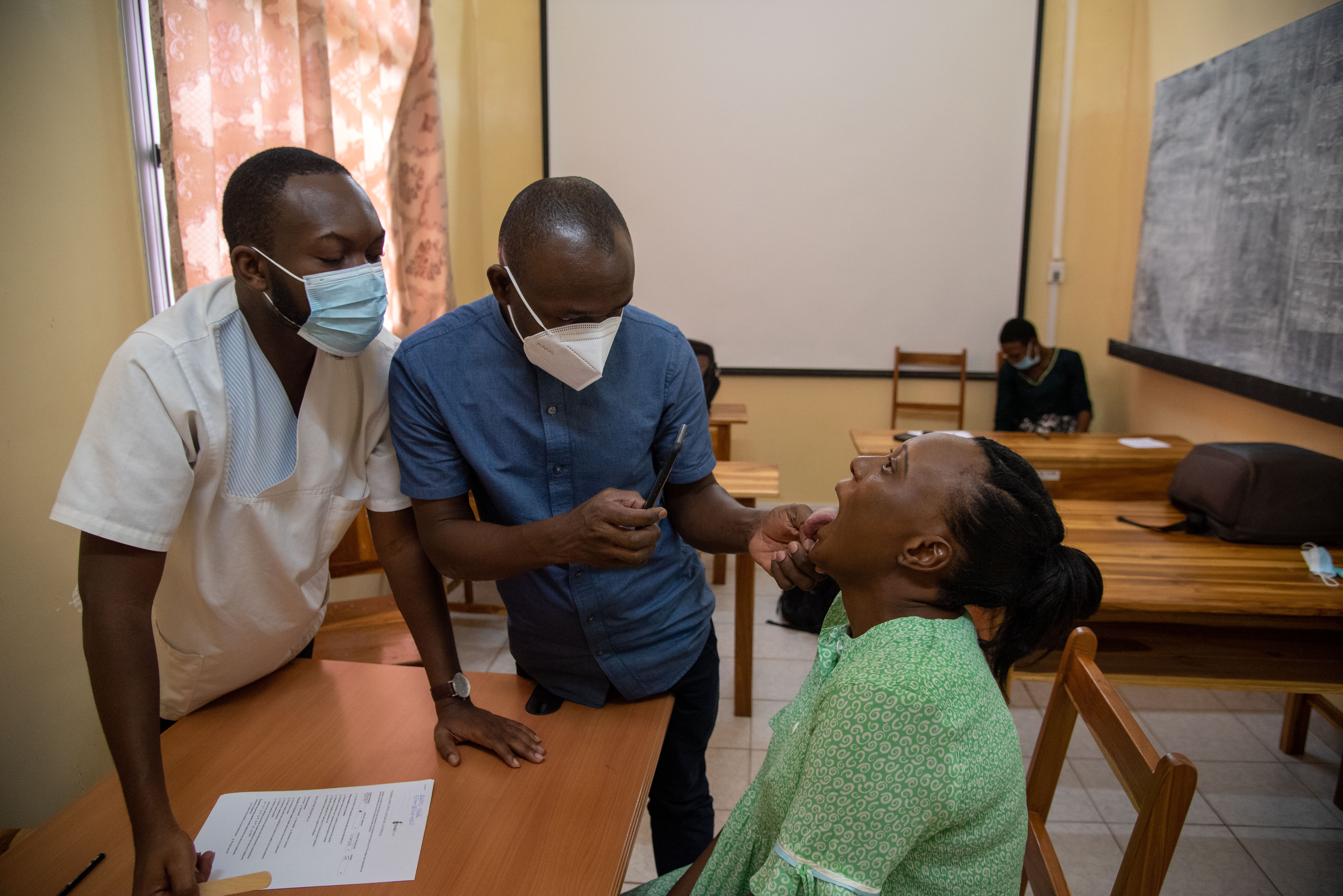 Edouardo taking a picture of the inside of an older patient’s mouth with her tongue sticking out. Faysal stands beside him. Both are wearing masks