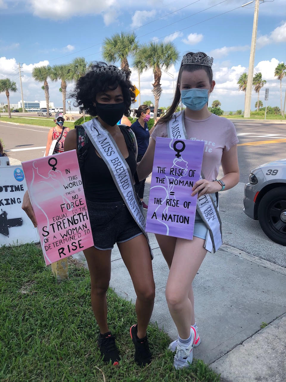 Alexandra and another woman wearing pageant sashes and masks holding women’s empowerment signs