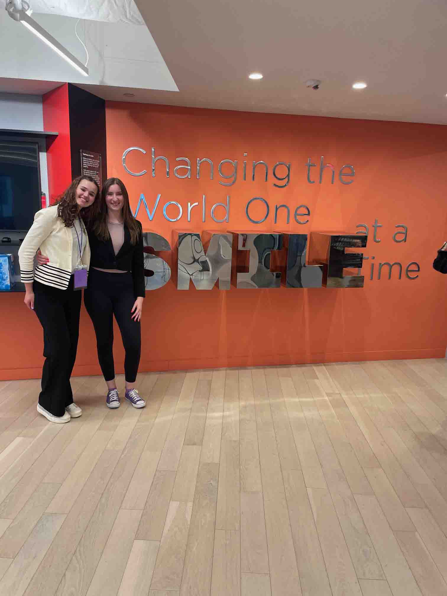 Alexandra and another girl standing in front of an orange wall in Smile Train’s office that says ‘Changing the world one smile at a time’