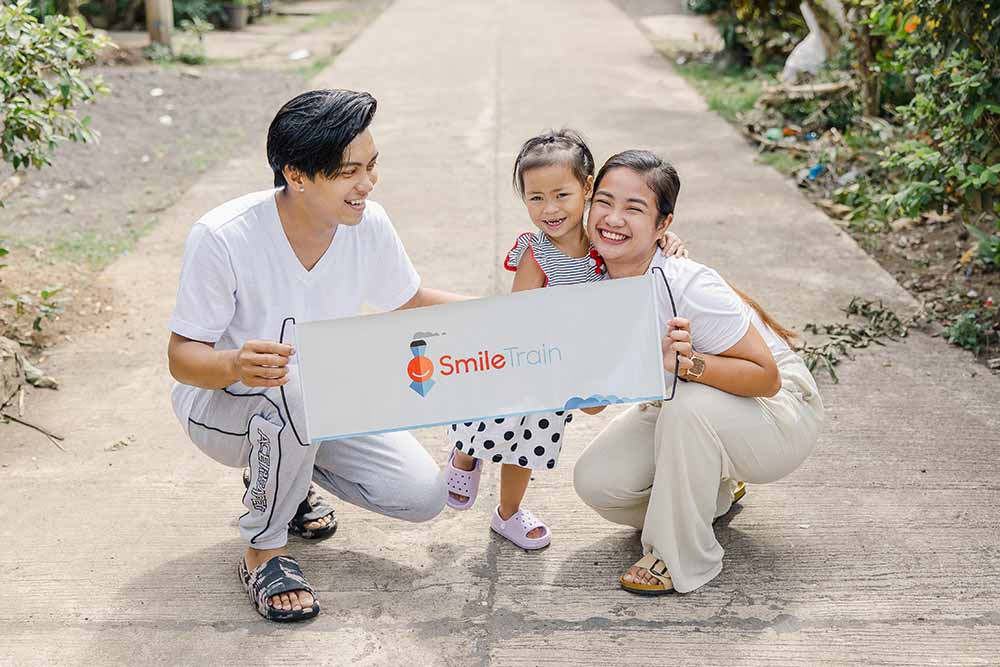 Ken, Kaylee, and Selma smile while holding up a Smile Train banner