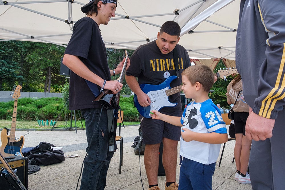 A child approaches a young man holding a guitar with a pick