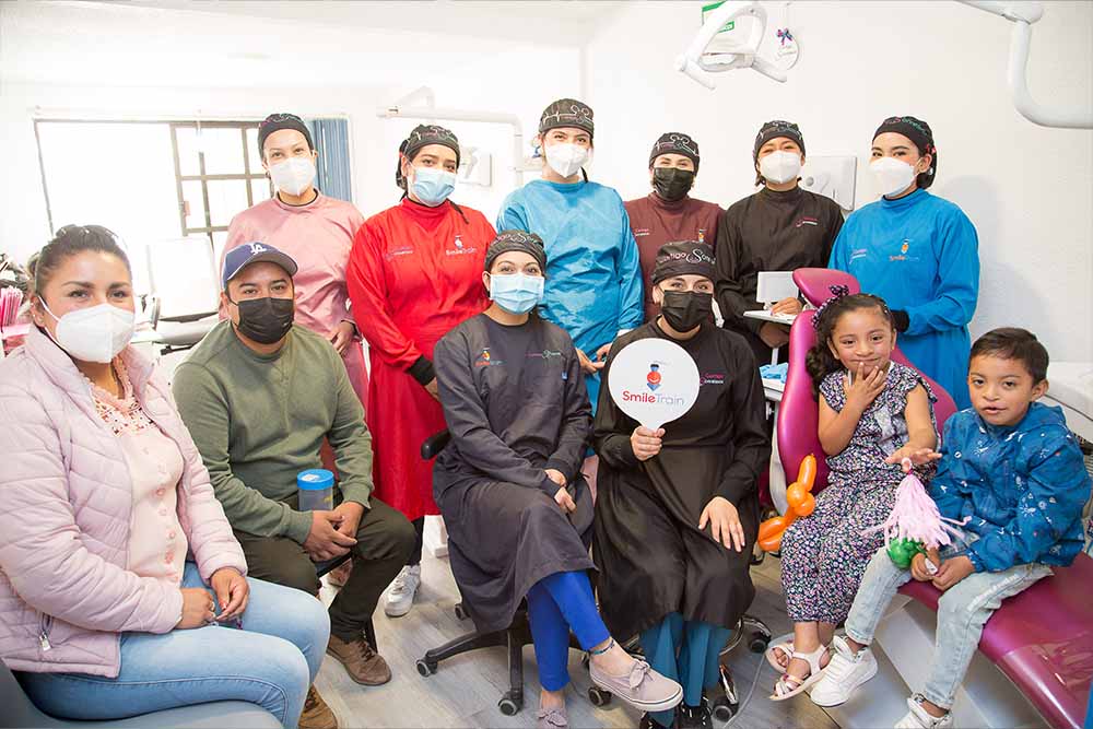 Leticia, Iván, and Ivanna sit along with another young cleft patient. In between them sit the staff of Contigo Sonreímos. Leticia, Iván, and the staff wear surgical masks
