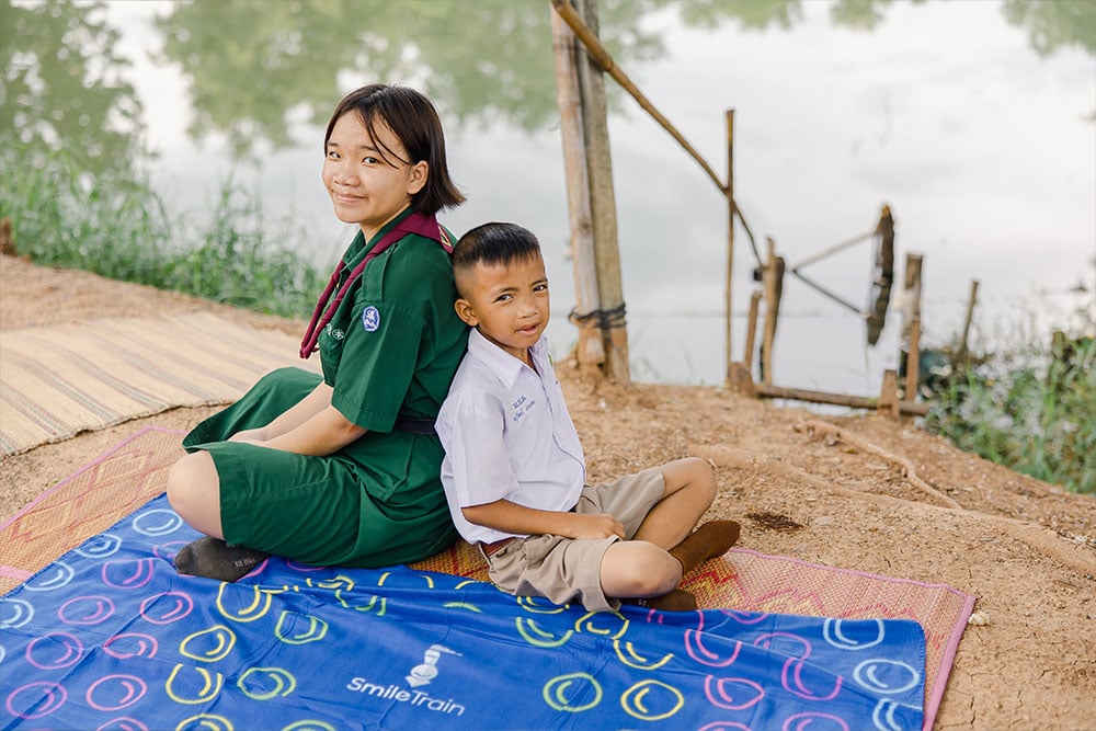 Anuwat and Aitya smiling, sitting back-to-back on the lakeshore