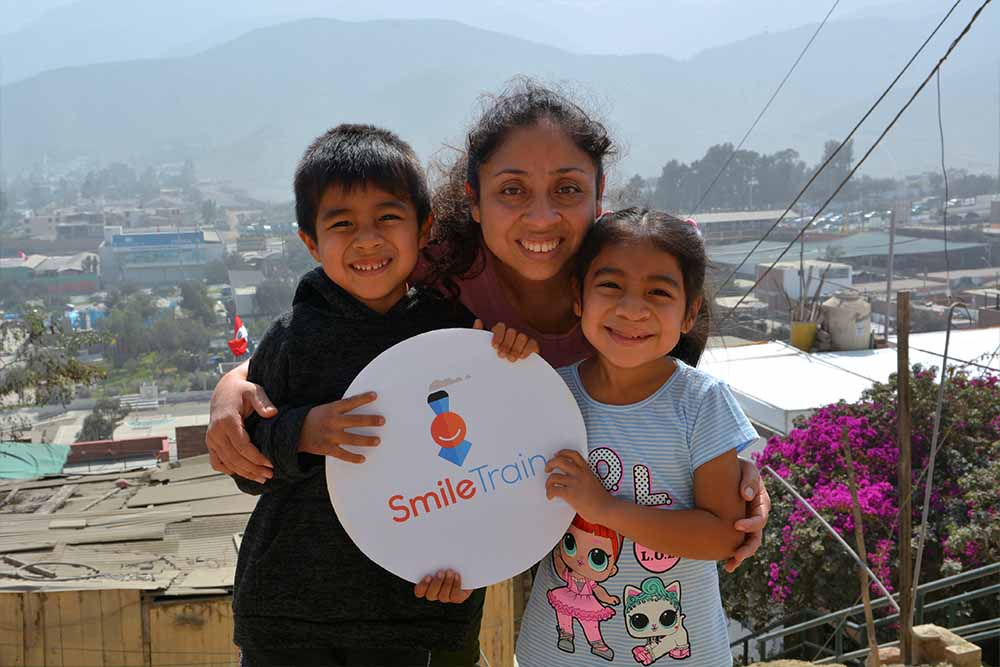 Outside in the sun, Astrid stands in a trio with her mother in the middle and her brother on the left. Together, they smile and hold up a small circular banner with Smile Train’s logo. Behind them stretches the terrain of Lima