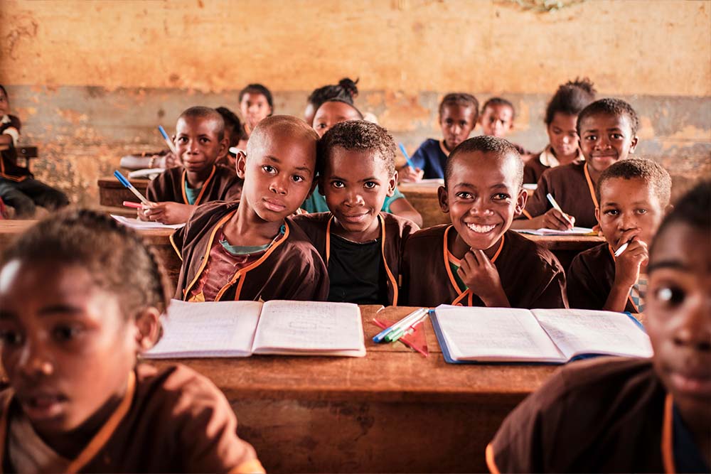 Ferdinand sits at his shared school desk with some friends. He wraps his arms around them, and together they all smile for the camera