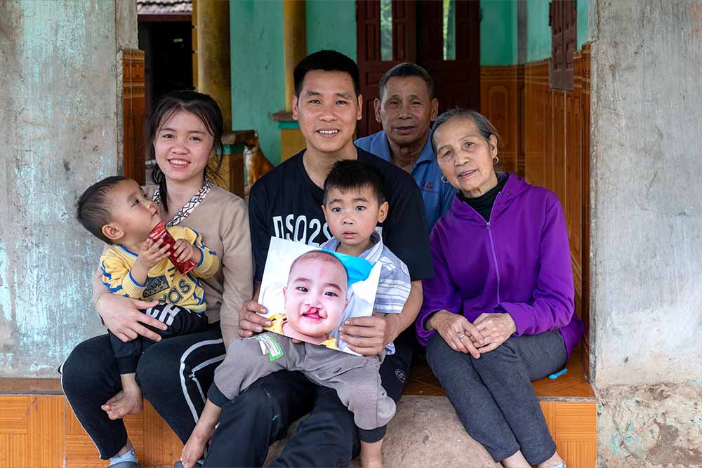 Gau sits in his father’s lap. Gau’s father holds up a photo of Gau from before cleft treatment. To their left sit Gau’s paternal grandparents. To their right sit Gau’s baby brother and Thuong