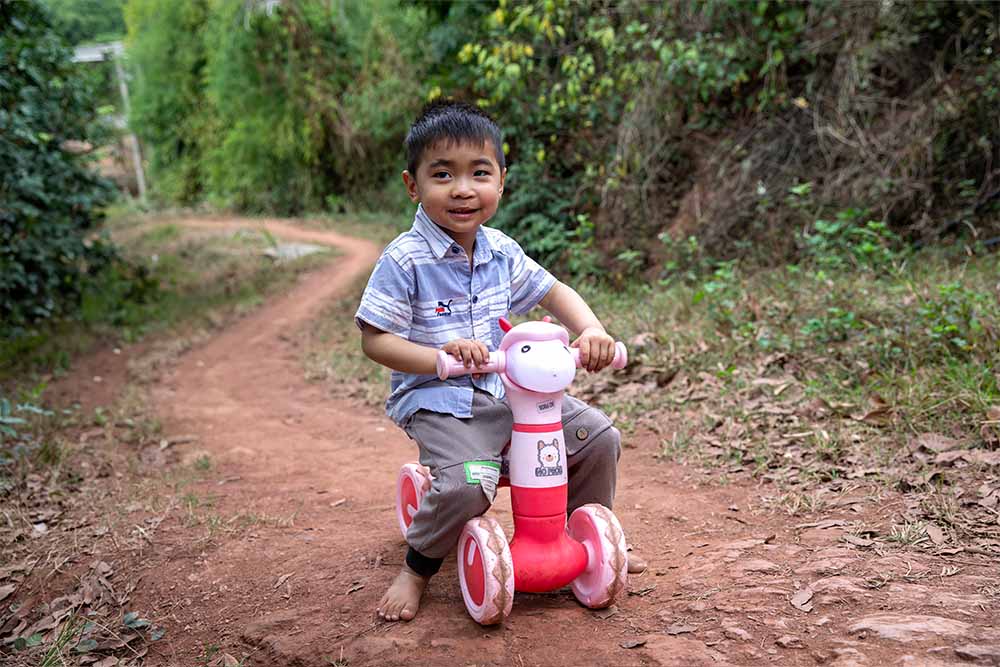 Gau smiles and sits on a small pink bike along a dirt path