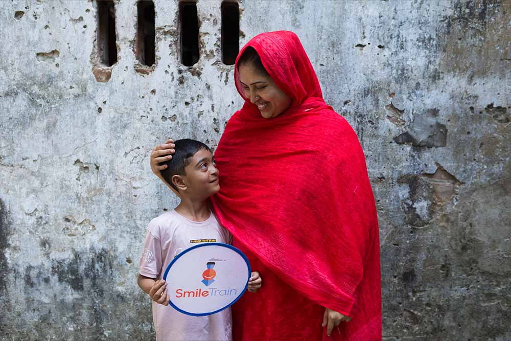 Holding a small, circular Smile Train banner, Maimun smiles and looks up at his mother, Muslima, who places a hand affectionately behind his head