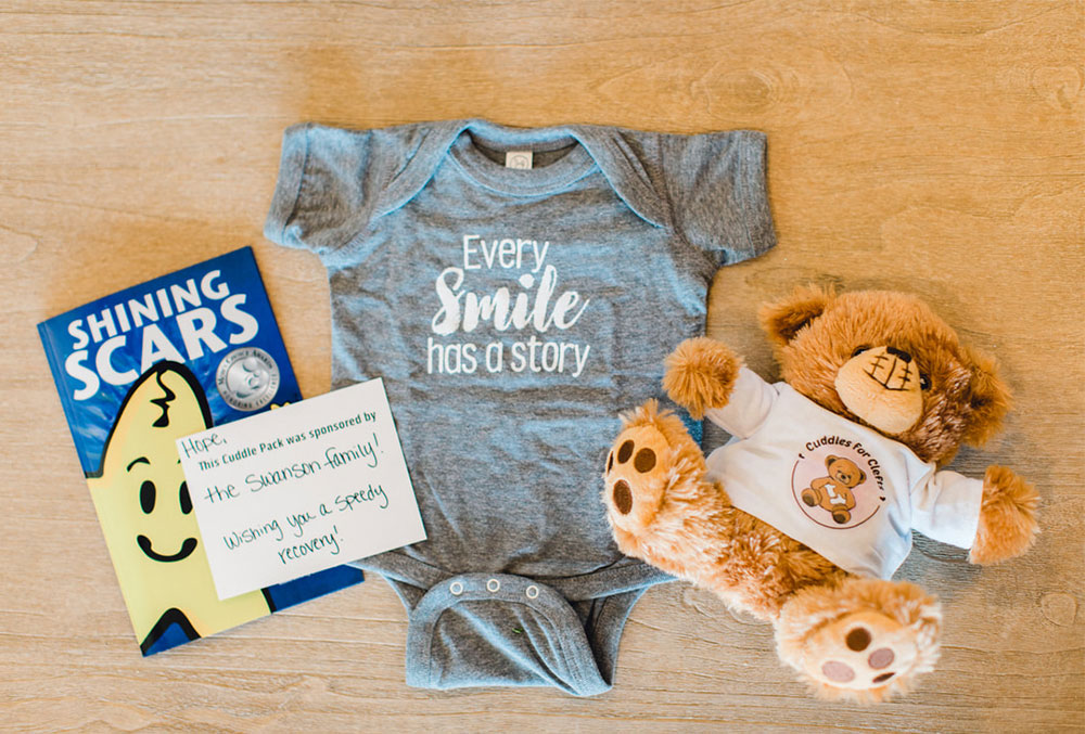 The contents of a Cuddle Pack: the book "Shining Scars" by Krystian Leonard, a 'Every Smile has a Story' onesie, a stuffed animal, and a note from the pack's sponsor