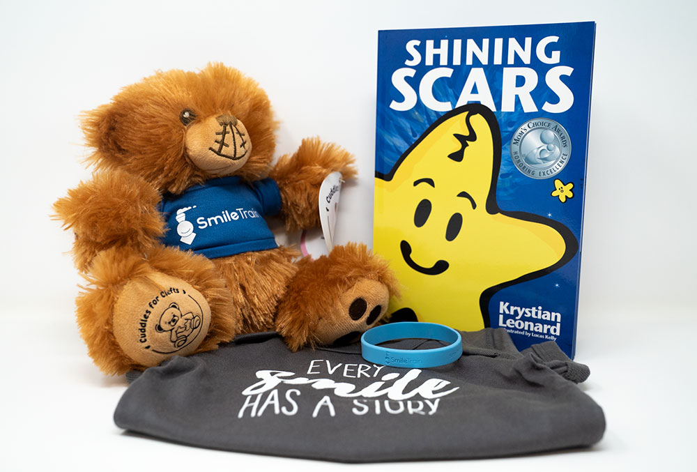 The contents of a Smile Train Cuddle Pack: a stuffed bear with a cleft wearing a Smile Train shirt, a 'Every Smile has a Story' onesie, a wristband, and the book "Shining Scars" by Krystian Leonard