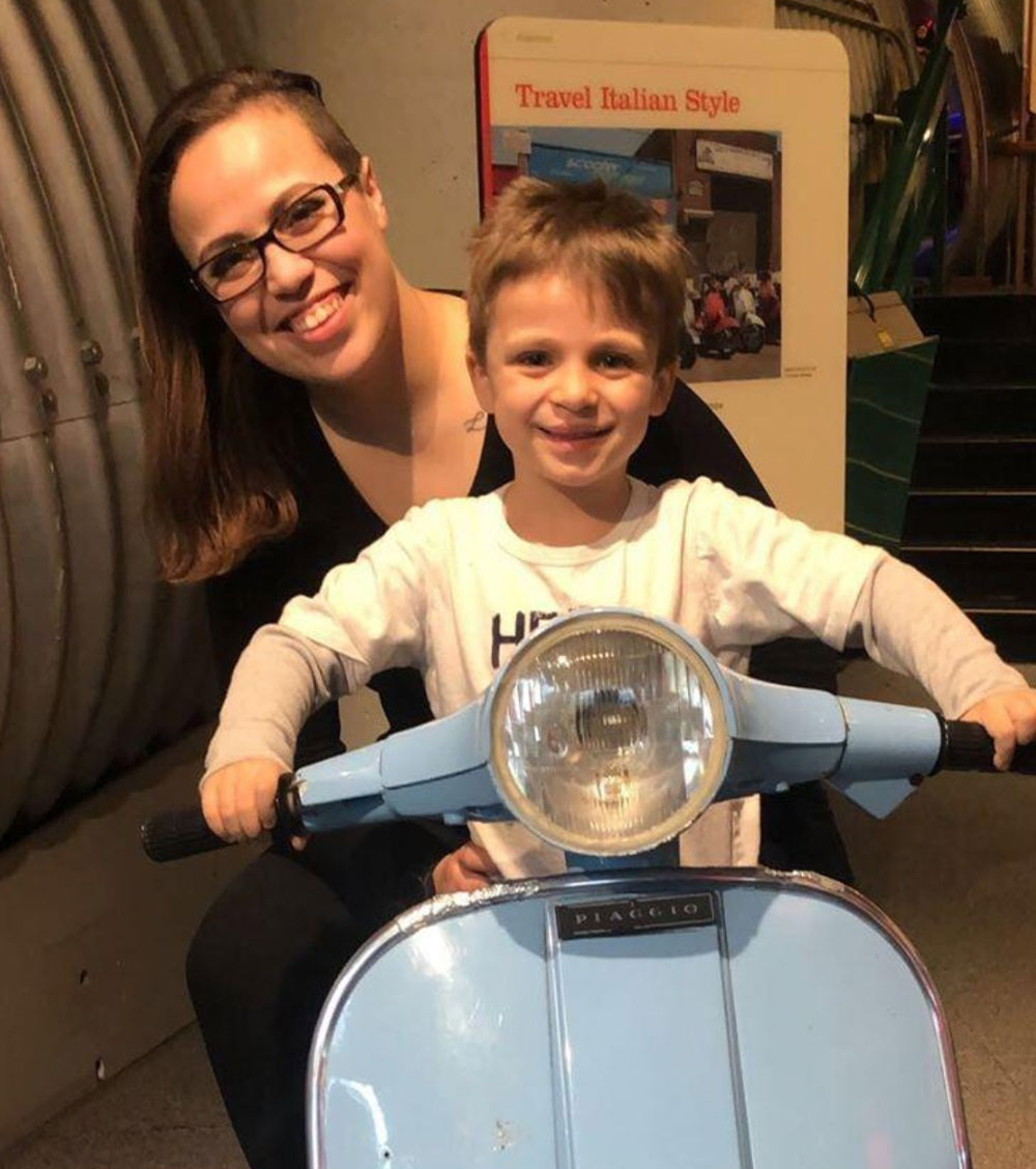 Jasmine and Daniel sit on a moped together, with Daniel pretending to drive.