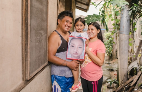 Savannah being held by her parents while holding a picture of herself before cleft lip and palate surgery. Everybody is smiling