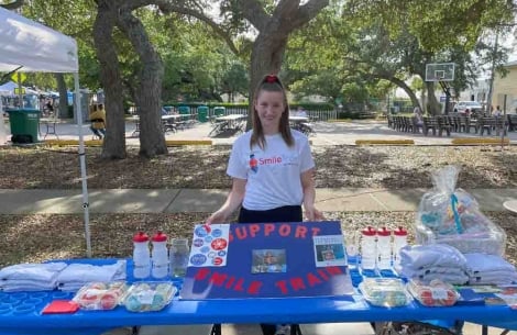Alexandra smiling and standing with a Smile Train support sign at a table