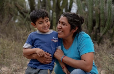 Walter smiles as Valeria kneels and wraps her arms around his affectionately. Behind them are shrubs and cacti”