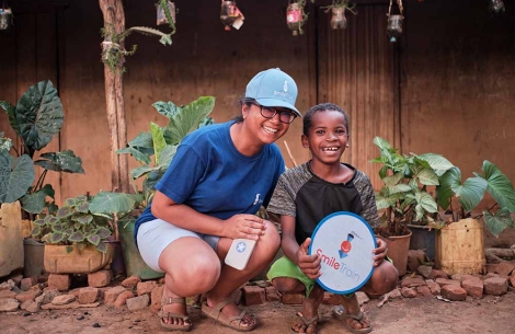 Ferdinand kneels and smiles with a Smile Train staff member. Ferdinand is holding a Smile Train circular logo.