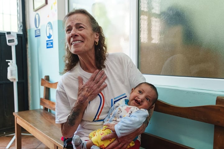 Karen Chambers holding a baby with a cleft with one arm, and her hand over her heart with emotion with the other