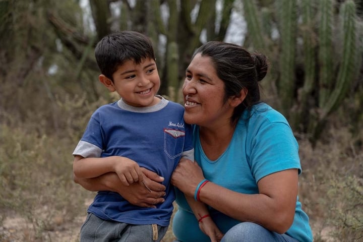 Walter smiles as Valeria kneels and wraps her arms around his affectionately. Behind them are shrubs and cacti”