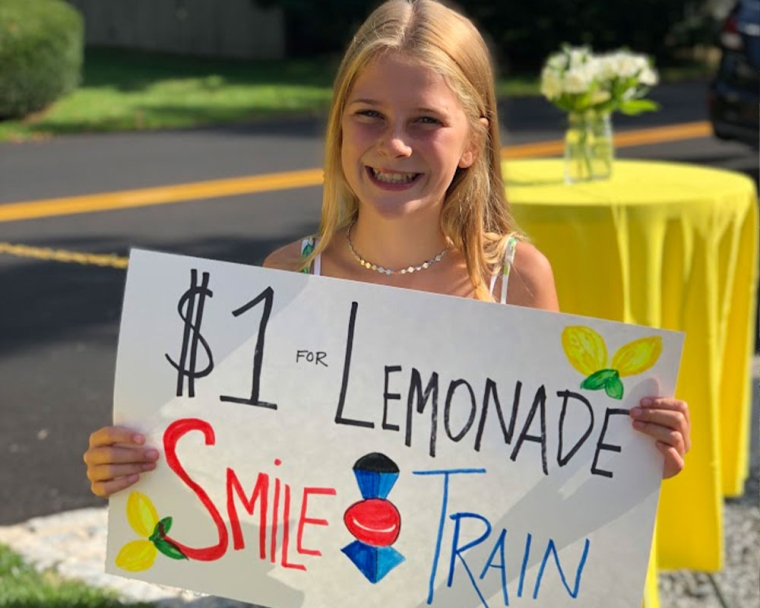Ella holding a sign that says "$1 for lemonade - Smile Train"