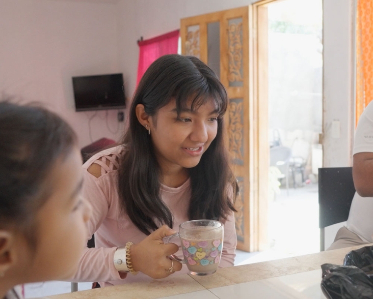 Adriana sits with her family at their kitchen table and enjoys a hot drink