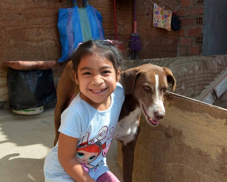 Outside their home, Astrid grins and kneels next to a lean brown-and-white dog, who’s also smiling.