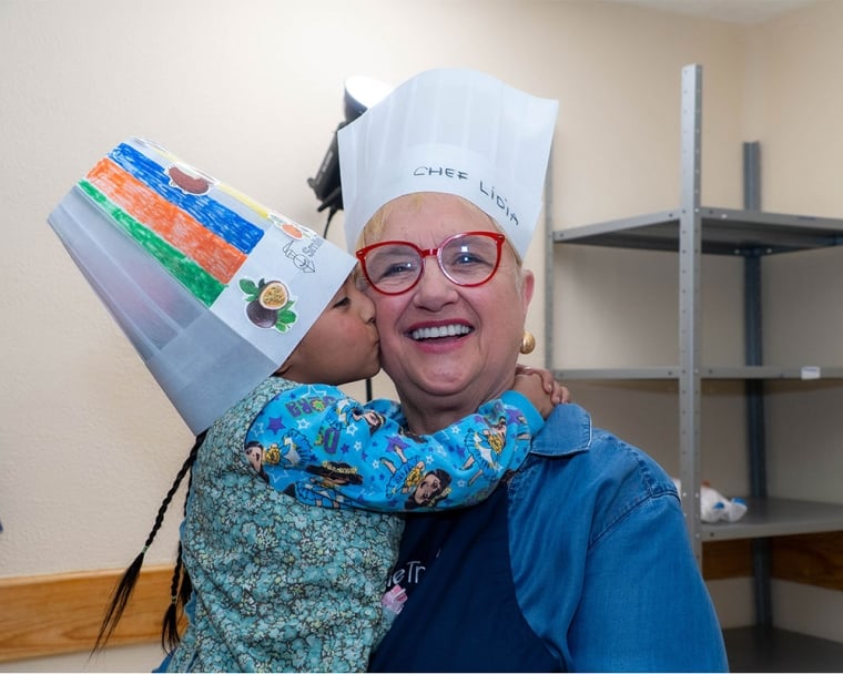 “Lidia holding a child in a hand-colored chef’s hat, who is kissing her cheek