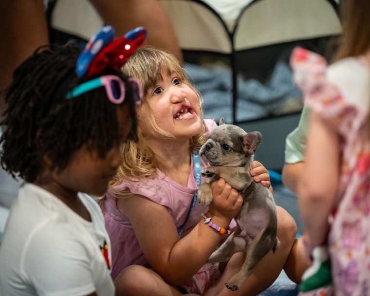 A little girl with a cleft smiles as she holds a puppy with a cleft